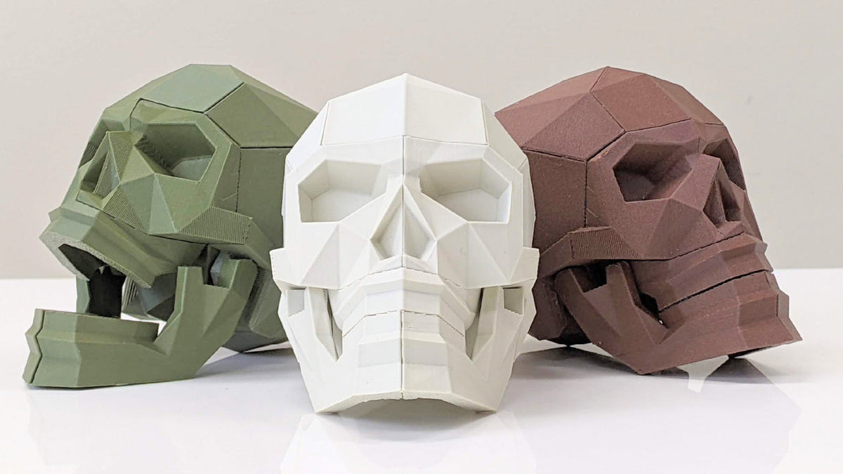 Three low-poly geometric skull models in olive green, white, and brown, displayed on a white reflective surface against a light gray background, with the Manabun Lab logo at the bottom.