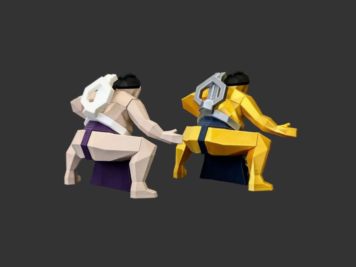 Two geometric, low-poly sumo wrestler figurines posed facing each other on a dark studio background. One figurine is beige with a purple waistband and black hair, and the other is yellow with a blue waistband and black hair.